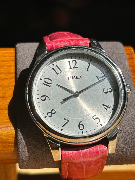 A 2013 Timex Quartz watch with a silver dial and pink leather strap shows 2:11, resting on a gray cushion on wood, bathed in sunlight.