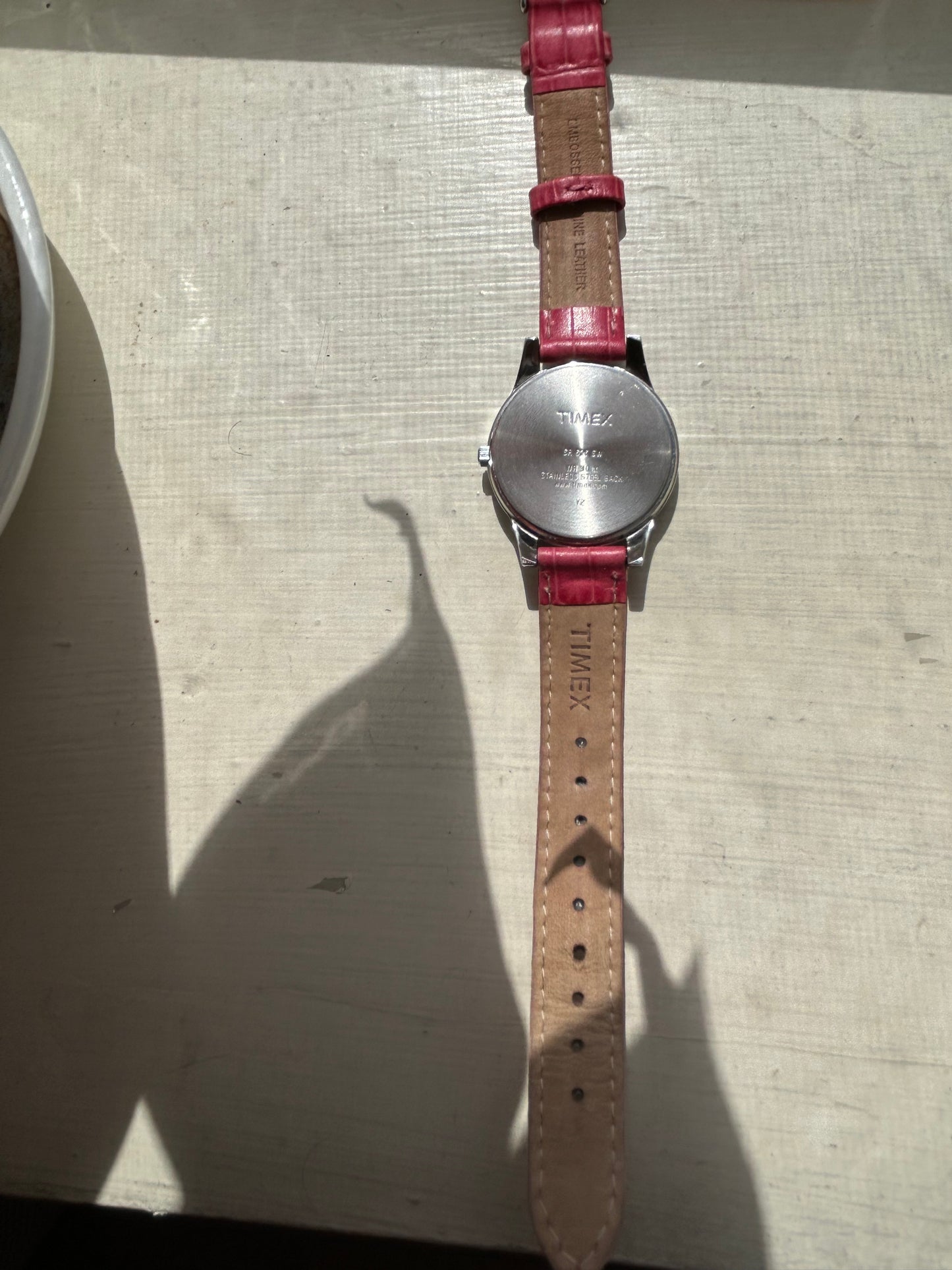 A 2013 Timex Quartz watch with a silver dial and pink leather strap lies face down on a light-colored surface, with sunlight casting a plant’s shadow beside it.