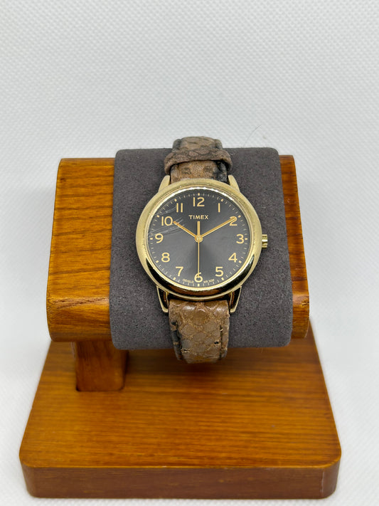 The 2012 Timex Black Dial Gold Case Snakeskin Strap Quartz Watch is displayed on a wooden stand with a gray cushion, set against a white textured background.