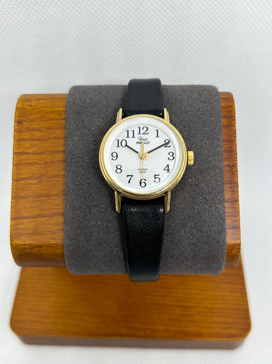 The Timex 1999 Gold Case Indiglo White Dial Quartz Watch with a black leather strap is displayed on a brown wooden stand with a gray padded holder against a white background.